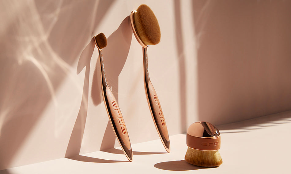 this is an image of 3 Elite Rose Gold brushes, two of which are leaning upright against the back wall and the third is sittin on the flat surface. Oval 7 Oval 3 and Palm Brush Mini