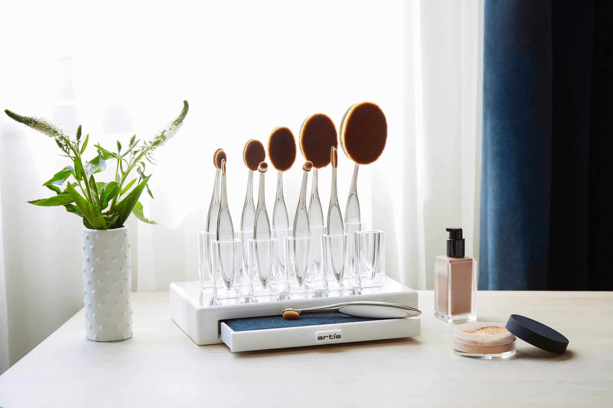 this is an image of a vanity room tabletop upon which is an Elite Collection Brush Holder Displyer unit with a drawer that slides out with a Brush Cleaning Pad. On the table is also a foundation product bottle and a jar of loose powder.